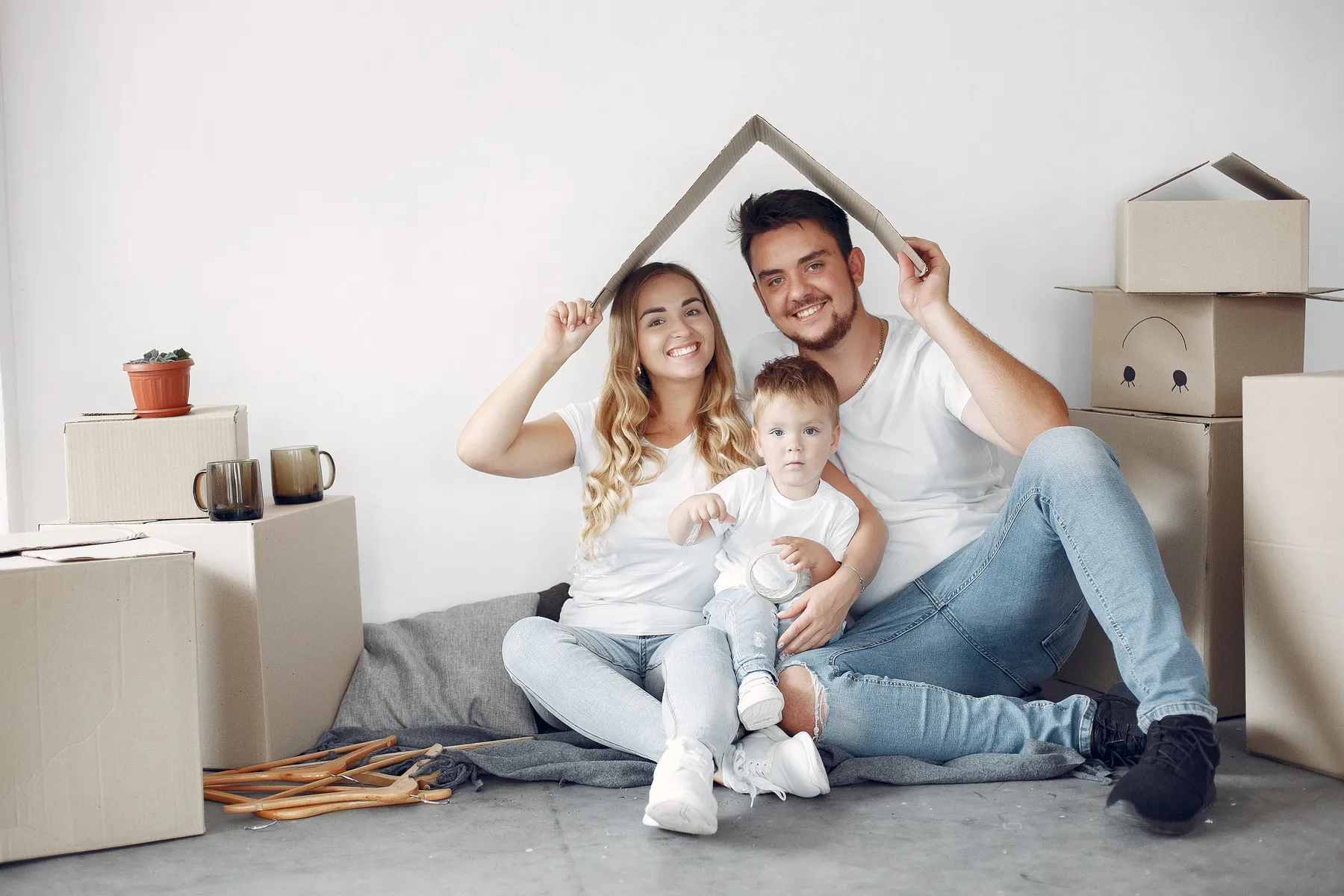 Familia joven sonriendo con un techo de cartón sobre sus cabezas, entre cajas de mudanza
